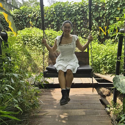 Hannah in a short white dress, sitting on a bench, smiling at the camera with lush green plants in the background.