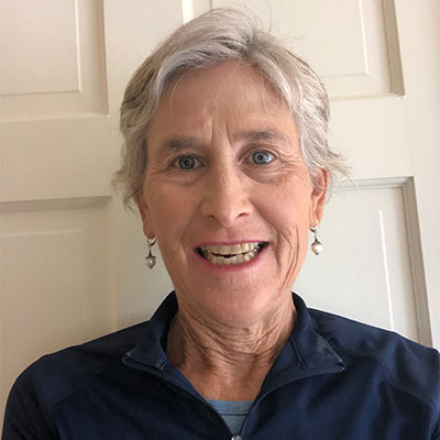 Julia with short gray hair, blue eyes, and earrings, smiling at the camera while standing in front of a cream-colored paneled door.