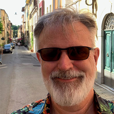 Kirk with gray hair, a beard, and sunglasses, smiling on a narrow street lined with buildings and parked cars.