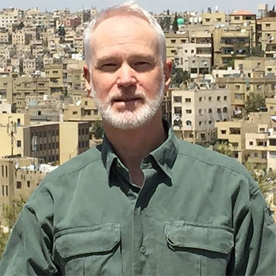 Rick with gray hair, a beard, in a green button-up shirt, looking at the camera with city apartment buildings on a hill in the background.